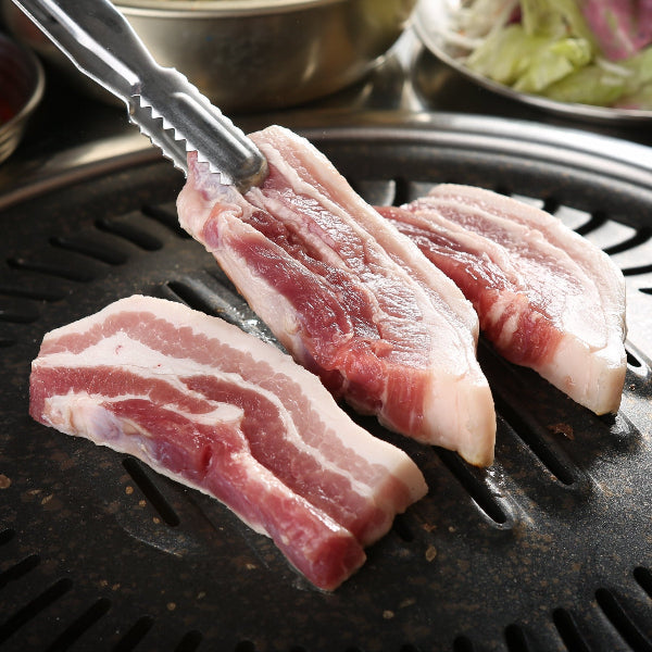 Raw pork belly being grilled on a hot plate with various side dishes in the background.