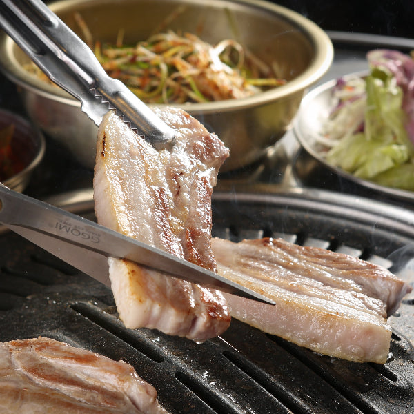 Bamboo skewers with grilled meat being held over a grill with various side dishes in the background.